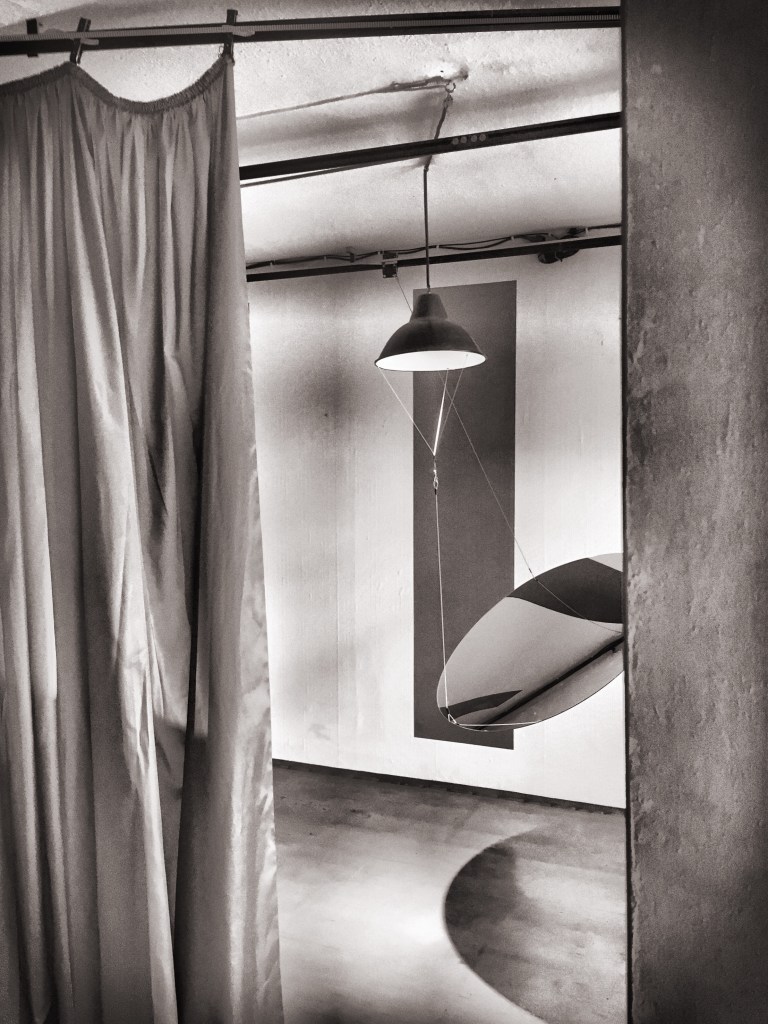 Ein Spiegel hängt in der Mitte eines Raumes. Rechts begrenzt eine Wand den Blick in den Raum mit dem Spiegel. Links begrenzt ein Vorhang den Blick. Eine Lampe hängt von der Zimmerdecke und beleuchtet den Spiegel. Das Bild ist schwarz/weiß.