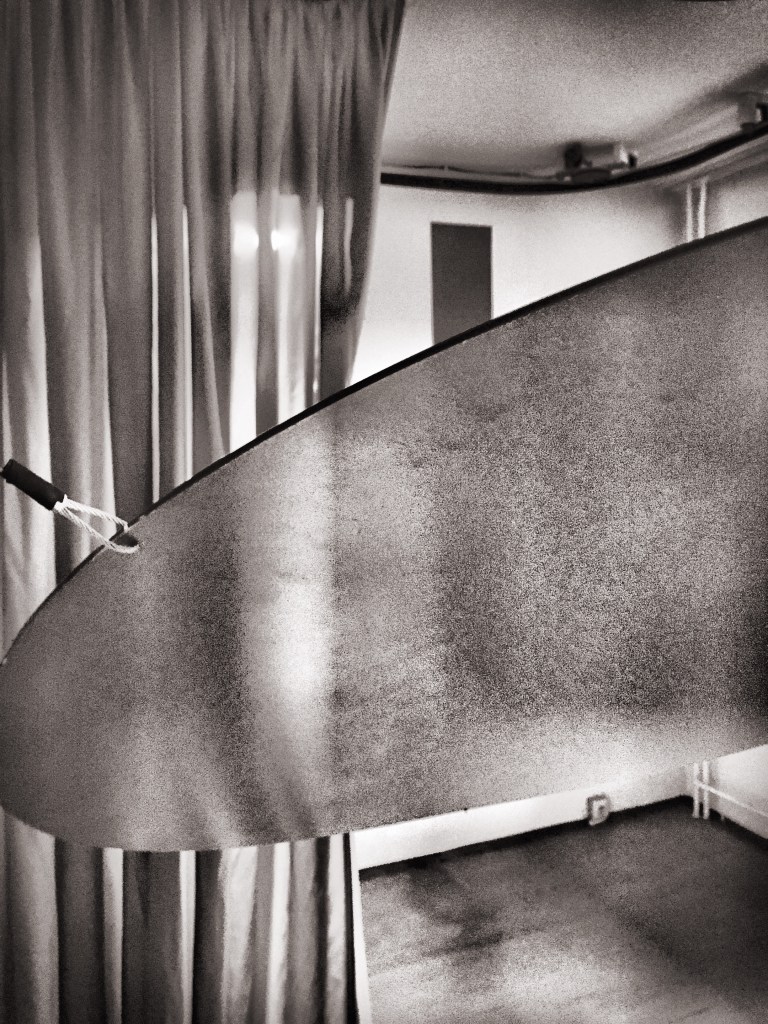 Nahaufnahme einer runden Platte, die in der Luft zu schweben scheint. An einer Ecke der Platte hängt ein Karabinerhaken. Im Hintergrund hängt ein Vorhang. Das Bild ist schwarz/weiß.