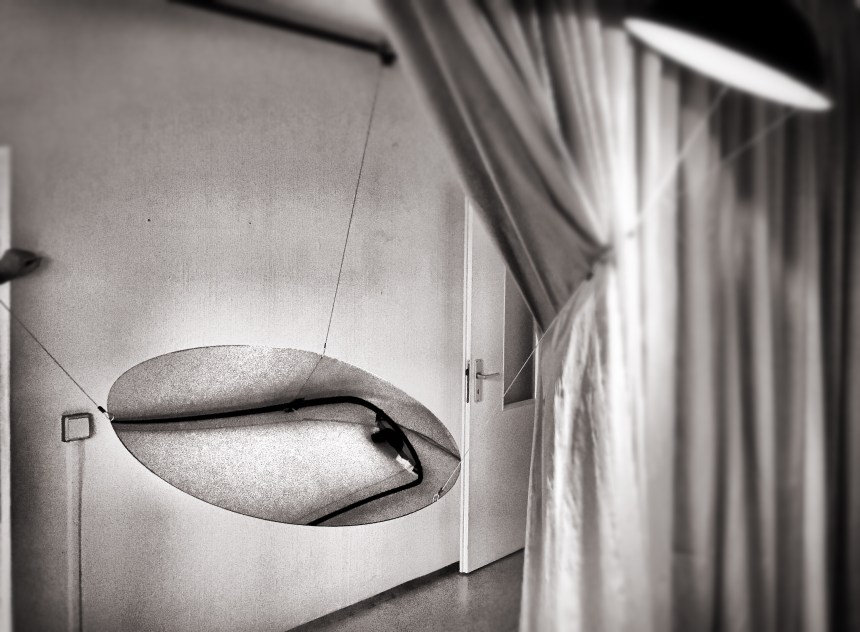 Ein Spiegel hängt in einem Raum an drei Metallseilen. Ein Vorhang schwebt im Bild. Ein Lampe hängt oben rechts und beleuchtet die Szenerie. Das Bild ist schwarz/weiß.