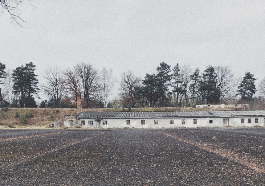 Ein breiter Schotterweg führt auf eine flache Baracke zu.An der linken Seite der Baracke steht ein hoher Schornstein. Dahinter ein Erdwall, der die Baracke leicht überragt. Auf dem Erdwall stehen Bäume.