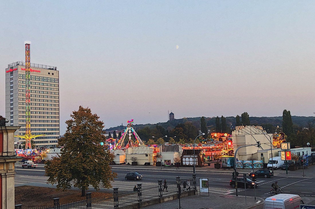 Volksfest auf einem Platz vor einem Hochhaus, Dämmerung, Lichter leuchten, ein Baum verliert langsam seine Blätter