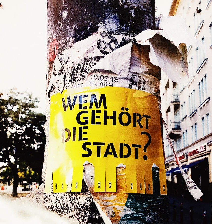 Ein Ausschnitt eines Laternenpfahls mit den Überresten von vielen Plakaten. Auf den Überresten klebt ein gelbes Plakat mit der Aufschrift "Wem gehört die Stadt?"
