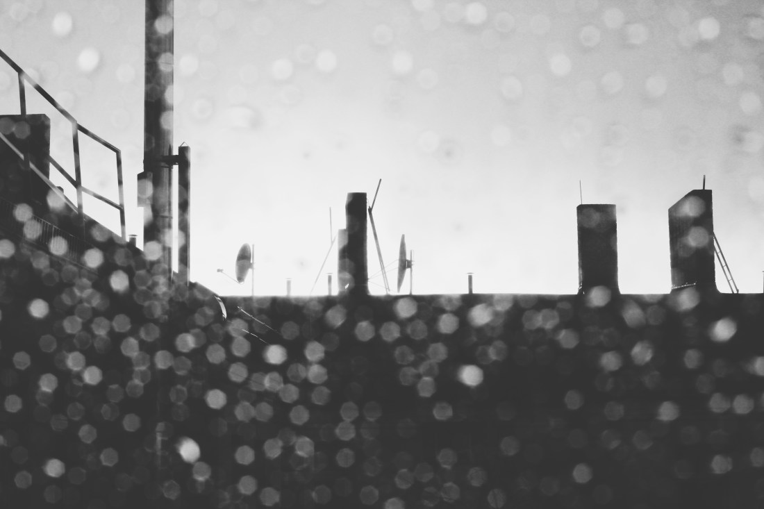 Schwarz/Weiß-Bild eines Dachs mit Schornsteinen und dem Sonnenuntergang. Im Vordergrund sind unscharfe Regentropfen auf dem Bild zu erkennen.