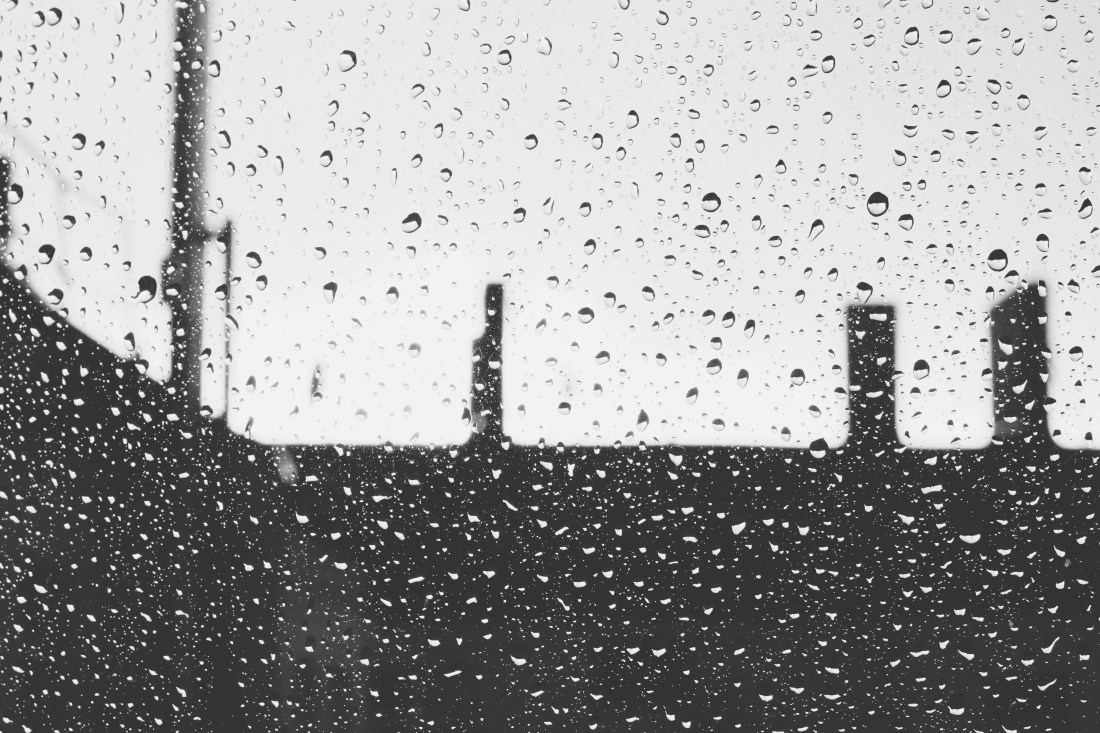 Schwarz/Weiß-Bild eines Dachs mit Schornstein und dem Sonnenuntergang. Der Hintergrund ist unscharf, im Vordergrund sind Regentropfen.