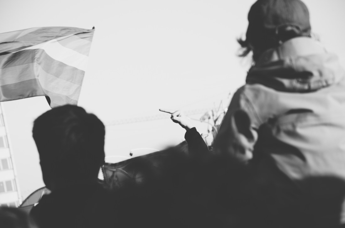 Eine unscharfe Menschenmenge im Vordergrund. Links wird eine Fahne geschwungen. Im Zentrum des Bildes zeigt jemand seinen Mittelfinger.