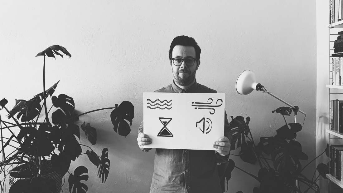 Schwarz-weiß Bild, ein Mann mit Brille hält ein Plakat auf dem ein Wellensymbol, ein Sturmsymbol, eine Sanduhr und ein Lautsprechersymbol zu sehen sind.