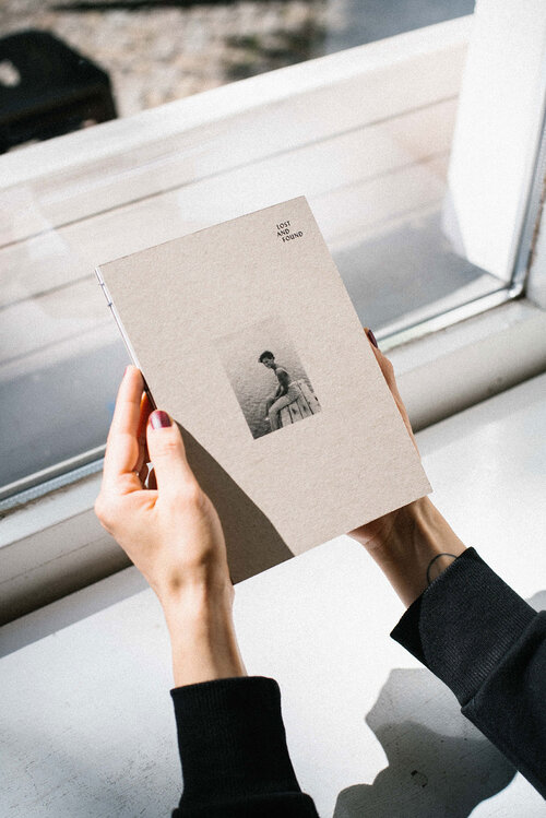 Zwei Hände halten das Buch Lost And Found vor einem Fenster in der Hand. Auf dem Cover des Buches ist ein Bild zu erkennen, darauf sitzt eine Frau auf einem Steg am Wasser.