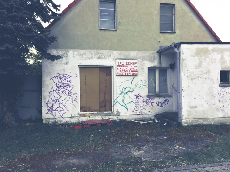 Altes, zweigeschossiges Haus mit flachem Anbau. Die Eingangstür fehlt und ist mit Spanplatten zugebaut. Das Sockelgeschoss ist weiß getüncht. Zwischen dem Fenster und der Eingangstür hängt ein Schild mit der Aufschrift "Tag Döner - Produktion".