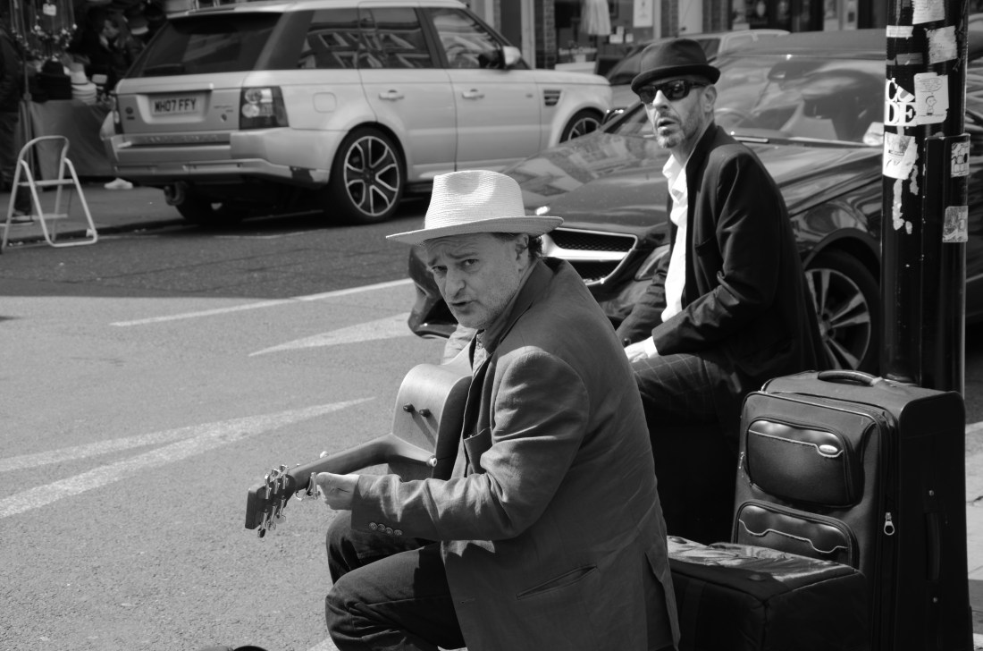 schwarz-weiß Bild, zwei Musiker sitzen an einer Straße, einer trägt einen weißen Hut mit Krempe, er spielt Gitarre und schaut skeptisch ins Bild. Der andere Musiker schaut aus dem Hintergrund ins Bild, erträgt einen schwarzen Hüten und Sonnenbrille. Zwischen den Musikern steht ein Koffer und ein Verstärker