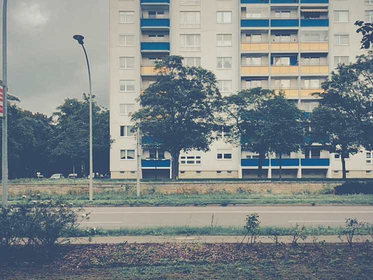 Im Vordergrund läuft eine Straße von rechts nach links. Ein Plattenbau steht im Hintergrund. Die verschieden farbigen Balkonbrüstungen sind blau und gelb. Eine Laterne steht links vor dem Haus.