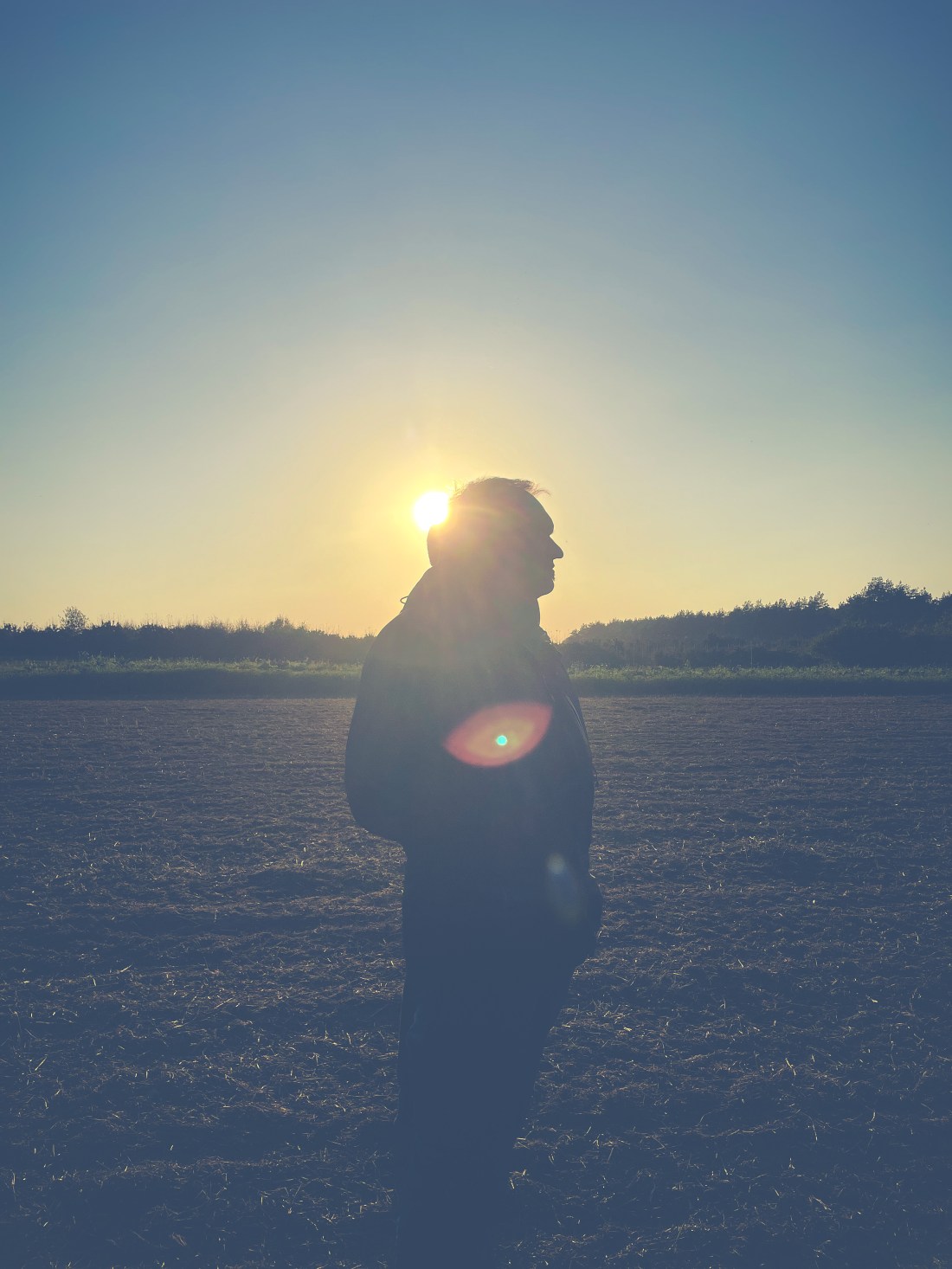 Ein Mann steht auf einem Feld, hinter ihm geht die Sonne langsam unter. Es ist nur sein Umriss zu erkennen.
