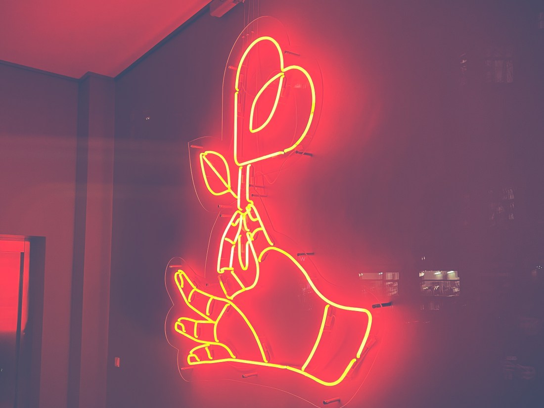 Eine Hand aus Leuchtröhren hält eine Blume, deren Blüte eine Herzform hat.