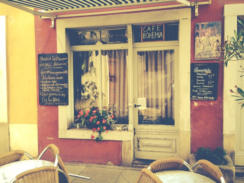 Eingang eines Cafés mit dem Namen Bohemia. Das Café ist geschlossen, davor stehen zwei Tische mit angelehnten Korbstühlen. Zwei Tafeln mit dem Angebot des Cafés hängen rechts und links vom Eingang.