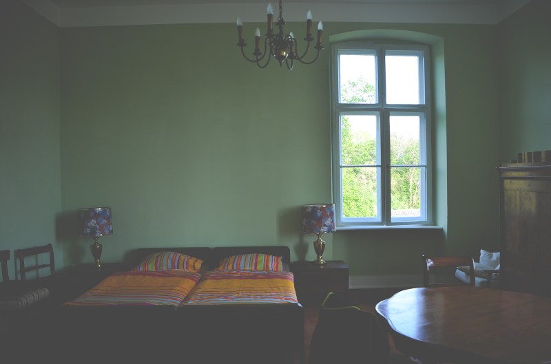 Zimmer mit grüner Wand, rechts ist ein Fenster, ein Schrank und ein Tisch. Links ist ein Bett mit zwei Lampen und bunter Bettwäsche.