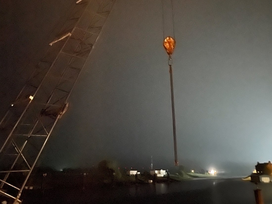 Fluss im Dunklen, von einem Kran hängt ein Seil herunter, ein Haken hängt am unteren Ende und eine lange Kette am Haken, in der Fern leuchten Boote am Ufer des Flusses