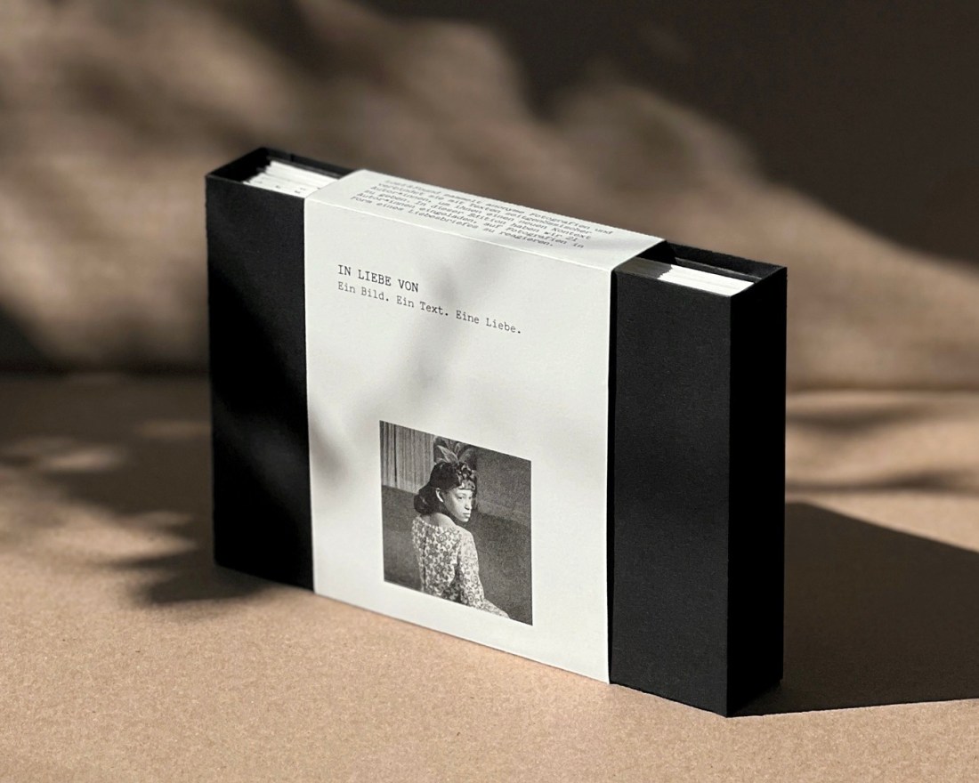 Schwarze Pappschachtel mit einer Banderole, darauf ist ein schwarz-ein-Bild einer Frau zu sehen, die über ihre rechte Schulter schaut. Über dem Foto steht der Schriftzug: IN LIEBE VON